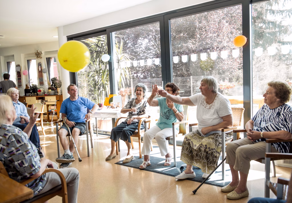 A group of elderly residents seated in a bright room with large windows, actively participating in a balloon game as part of the care home lifestyle at Oakham